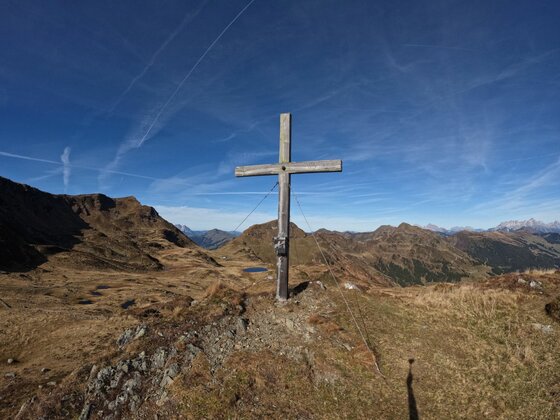 Ein hölzernes Gipfelkreuz auf einem grasbewachsenen Bergrücken vor einem Panorama aus braunen Alpengipfeln.