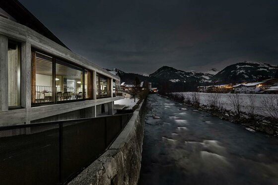 Modernes Gebäude mit großen Glasfronten bei Nacht neben einem Fluss in winterlicher Berglandschaft.
