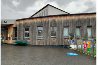 Ein modernes Gebäude mit Holzfassade und großen Fenstern. Vor dem Eingang sind Sitzgelegenheiten und ein Parkplatz für Menschen mit Behinderung.