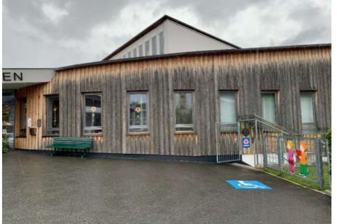 Ein modernes Gebäude mit Holzfassade und großen Fenstern. Vor dem Eingang sind Sitzgelegenheiten und ein Parkplatz für Menschen mit Behinderung.