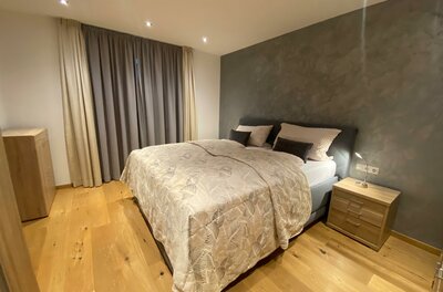 Ein modernes Schlafzimmer mit einem Doppelbett und Holzfußboden. Die Wände sind in neutralen Farben gehalten, und es gibt Vorhänge sowie eine Kommode.