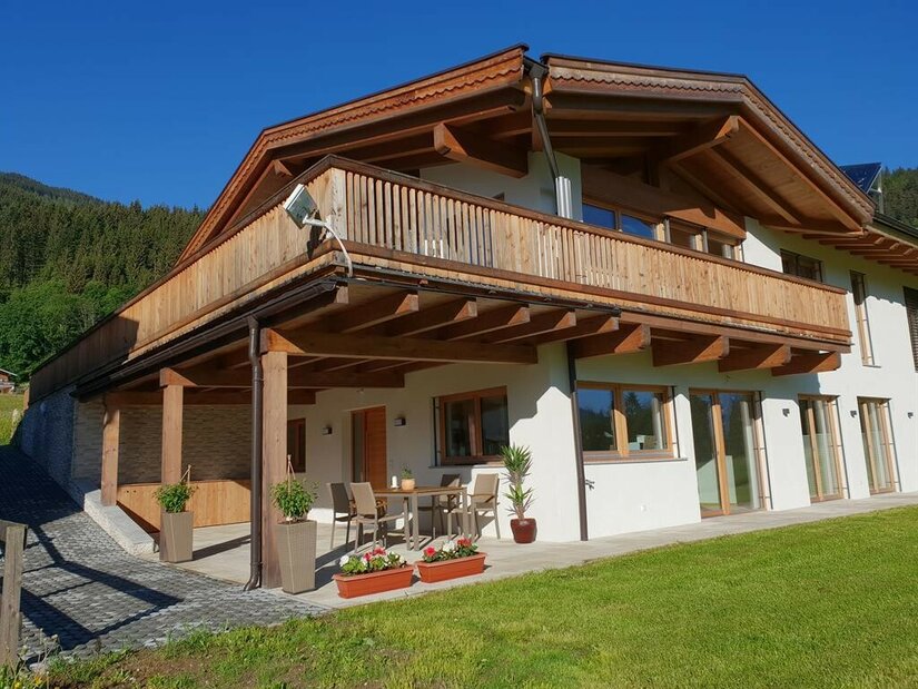 Zweistöckiges alpines Wohnhaus mit Holzfassade, Balkon und Terrasse vor einer Wiese.