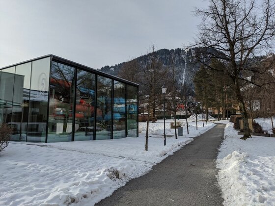 Glasgebäude an Winterwanderweg Modernes Glasgebäude neben einem geräumten Fußweg in einer verschneiten Parkanlage vor bewaldetem Berg.