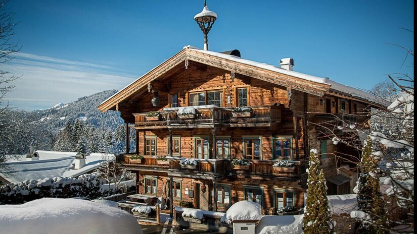 Mehrstöckiges Holzhaus mit Balkonen und schneebedecktem Dach vor bewaldeten Bergen im Winter.