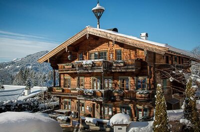 Mehrstöckiges Holzhaus mit Balkonen und schneebedecktem Dach vor bewaldeten Bergen im Winter.