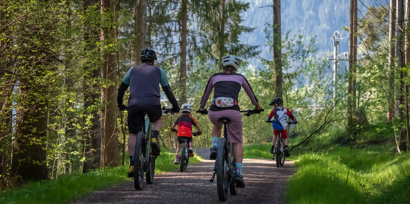 Eine Familie fährt mit Fahrrädern auf einem schmalen Waldweg, umgeben von frischem Grün und hohen Bäumen.