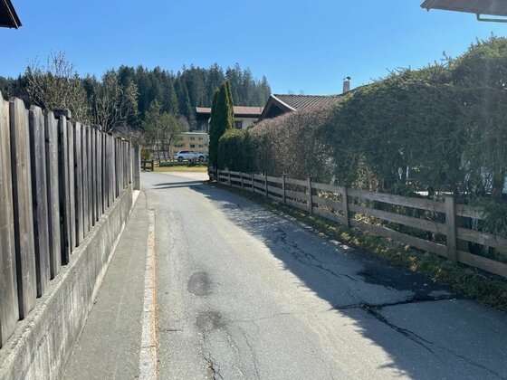 Asphaltierter Weg zwischen Holzzäunen und Hecken in einer Siedlung vor bewaldetem Hintergrund.