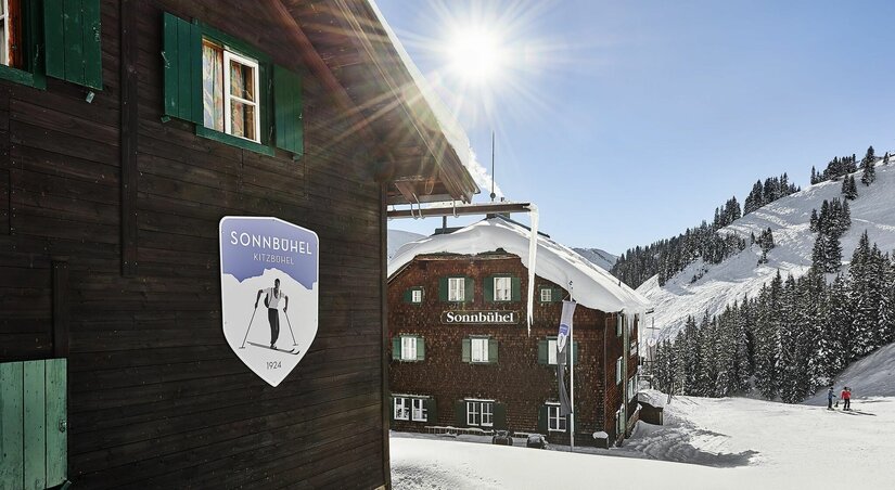 Holzhütte Sonnbühel im Schnee mit Logo an der Fassade und verschneiten Bergen im Hintergrund bei Sonnenschein.