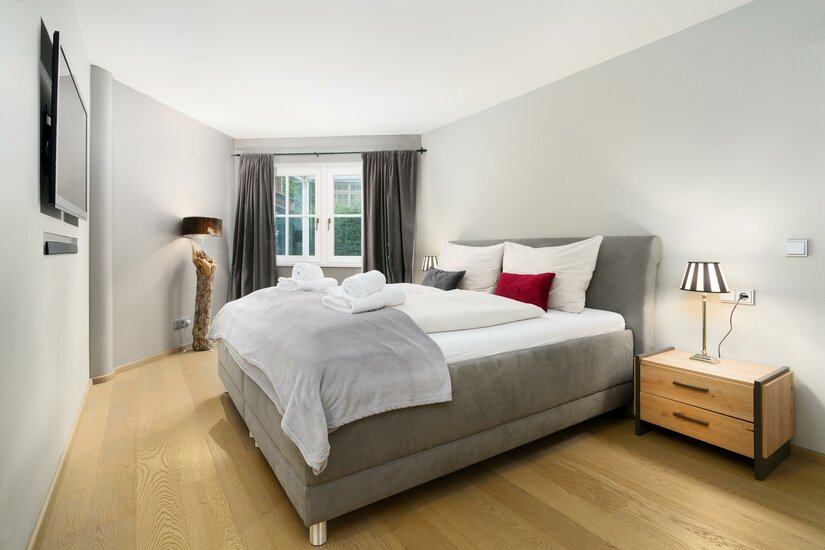 Modernes Schlafzimmer mit grauem Polsterbett, Holzparkett, Nachttisch und Fenster mit dunklen Vorhängen.