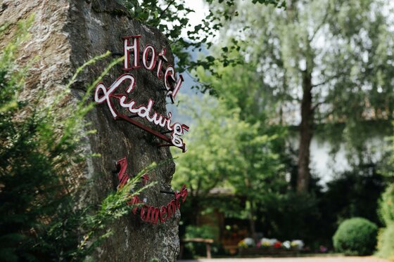 Ein großer Naturstein mit dem roten Schriftzug Hotel Ludwig vor grünen Bäumen.