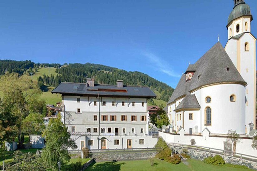 Schwarzer Adler im Sommer Mehrstöckiges weißes Gebäude neben einer Kirche mit Zwiebelturm vor bewaldeten Bergen.