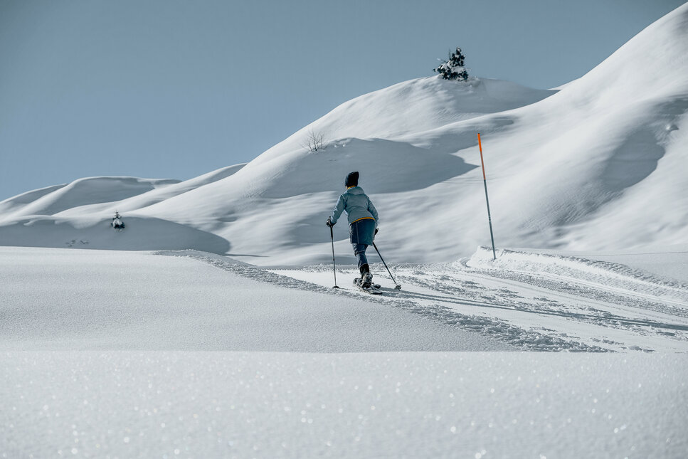 Eine Person wandert durch eine schneebedeckte Landschaft, umgeben von sanften Hügeln und klarem blauen Himmel.