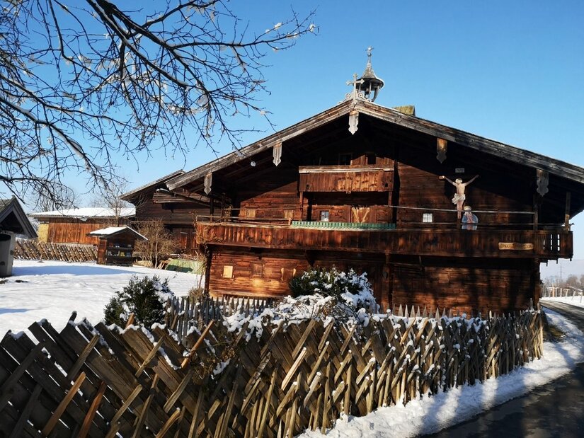 Traditionelles Bauernhaus aus dunklem Holz mit Holzzaun im Schnee unter blauem Himmel.