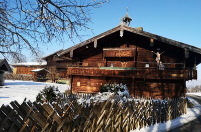 Traditionelles Bauernhaus aus dunklem Holz mit Holzzaun im Schnee unter blauem Himmel.