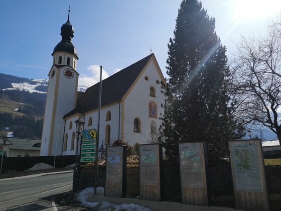 Weiße Kirche mit Zwiebelturm vor bewaldeten Bergen und Informationstafeln im Vordergrund.