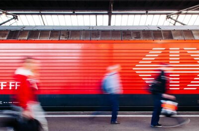 Seitenansicht eines roten Zuges am Bahnsteig mit unscharf vorbeilaufenden Personen.