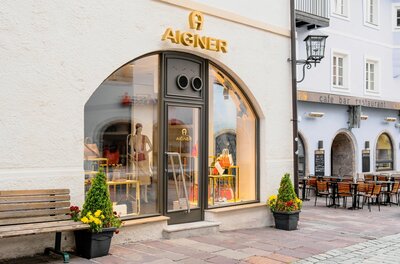 Außenansicht des Aigner Shops mit Schaufenster, Taschen und einer Parkbank davor.