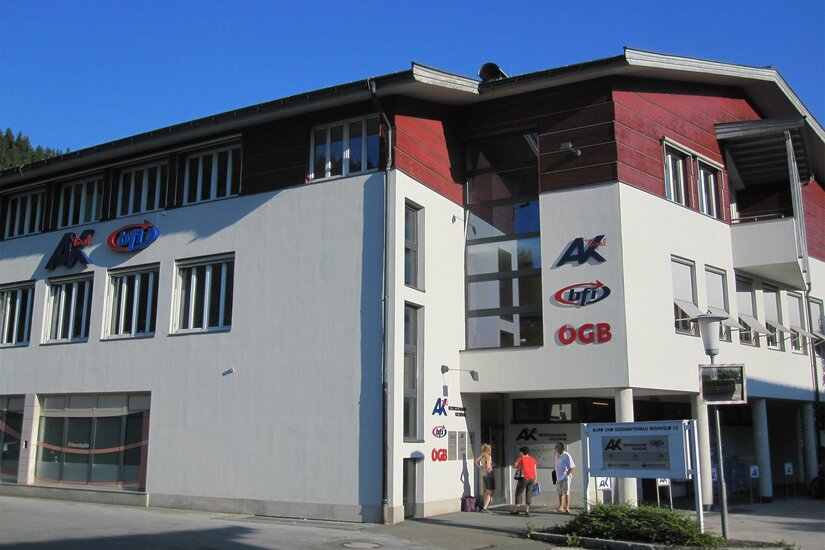 BFI Kitzbühel Ein modernes Gebäude mit dem Logo der AK und OGB. Es ist klarer Himmel und sonniges Wetter zu sehen.