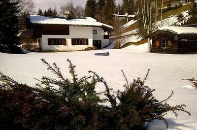 Haus Winter Alpines Wohnhaus mit schneebedecktem Dach und kleiner Holzhütte auf einer verschneiten Wiese.