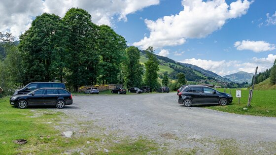 Parkplatz Schradlern Ein geschotterter Parkplatz mit mehreren Autos vor einer Kulisse aus grünen Bäumen und Bergen.