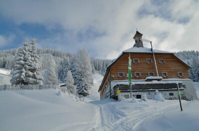 Holzhütte mit Glockenturm in verschneiter Winterlandschaft vor bewaldetem Hang.