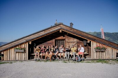 Eine Gruppe von Wanderern sitzt mit einem Hund auf einer Holzbank vor einer rustikalen Almhütte unter blauem Himmel.