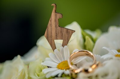 <p>Eine Holzfigur in Form einer Gams steht auf einer bunten Blumenarrangierung. Im Vordergrund sind goldene Ringe zusammen mit weißen Margeriten zu sehen.</p>