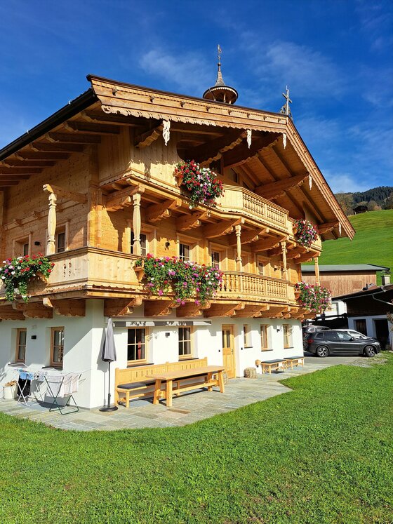 Ein traditionelles Chalet mit Holzverkleidung und Balkonen, geschmückt mit bunten Blumen. Im Hintergrund sieht man grüne Wiesen und einen blauen Himmel.