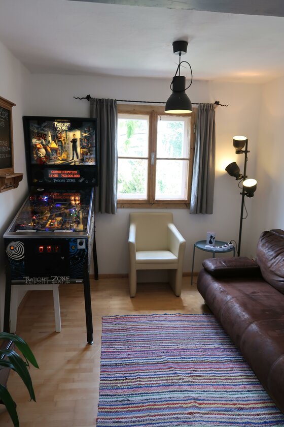 Innenansicht eines Zimmers mit Flipperautomat, Ledersofa und hellem Sessel vor einem Fenster.