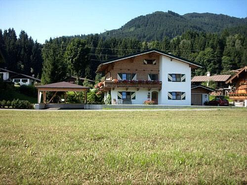 Haus Mitterer im Sommer Alpines Wohnhaus mit weißer Fassade, Holzbalkon und Fensterläden vor bewaldetem Berg.
