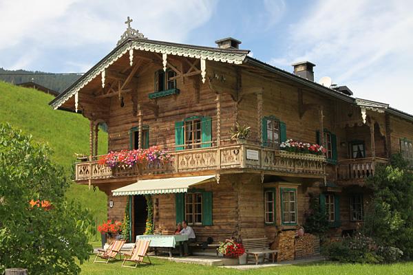 Landhaus Kainzern im Sommer Zweistöckiges Holzhaus im alpinen Stil mit Blumenbalkonen und grünen Fensterläden vor einer Wiese.