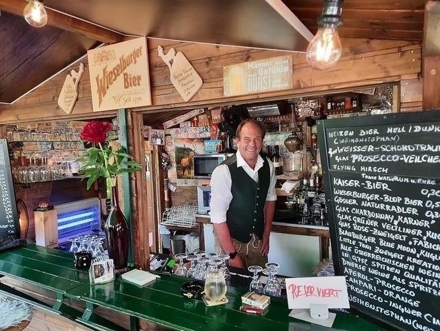 Fritz' LICHTBLICK Ausschank Ein Mann in Trachtenweste steht hinter einem hölzernen Tresen mit Getränkekarte und Gläsern.