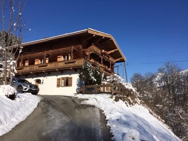 Ferienhaus Giggling 3 im Winter Zweistöckiges Holzhaus mit weißem Erdgeschoss in verschneiter Berglandschaft unter blauem Himmel.