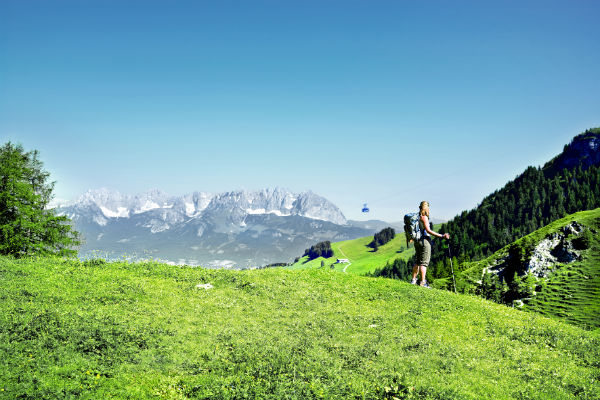 Wanderin mit Rucksack auf einer grünen Bergwiese vor einem Gebirgspanorama mit Seilbahngondel.