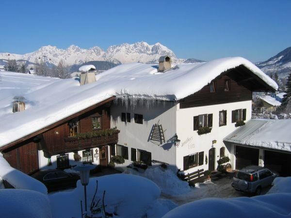 Landhaus Eva im Winter Schneebedecktes Landhaus mit dunkler Holzfassade vor einem Gebirgspanorama unter blauem Himmel.
