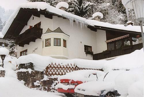 Haus Schatz im Winter Schneebedecktes alpines Wohnhaus mit Eiszapfen am Dach und eingeschneiten Autos im Vordergrund.