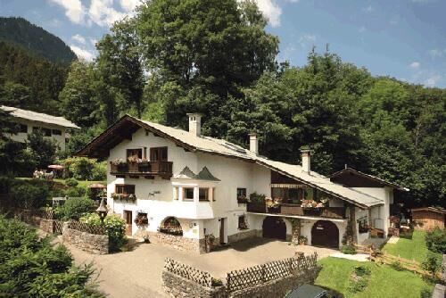 Haus Schatz im Sommer Zweistöckiges weißes Alpenhaus mit Holzbalkonen und Blumenkästen vor bewaldetem Hang.